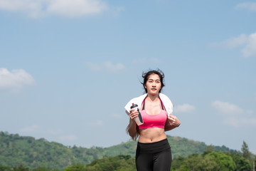 Front view Young asian woman running on road in the morning sunshine outdoors. Female jogger exercising in nature outdoors holding bottled water. Fitness and healthy lifestyle