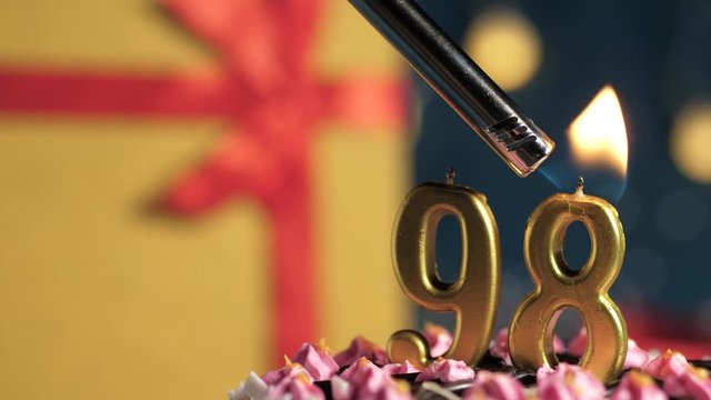 Birthday cake number 98 golden candles burning by lighter, blue background gift yellow box tied up with red ribbon. Close-up and slow motion