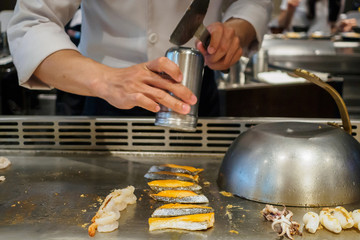 Japanese chef cooking and sprinkling spices on seafood in teppanyaki restaurant.