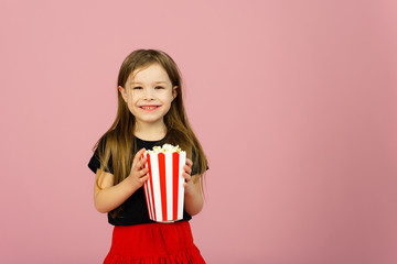 Happy cute little girl holding popcorn in her hands and looking at the camera on a pink background. The concept of watching a movie in a movie theater or at home. Copyspace - obrazy, fototapety, plakaty