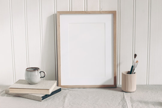 Blank wooden frame mockup with paint brushes, pencils in ceramic holder, cup of coffee and books on linen tablecloth. Artistic scene. Creative table background. Poster product design, wooden wall.