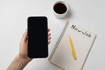 Female hand holding phone. Notepad, pen and cup of coffee on white background. New job. Copy space