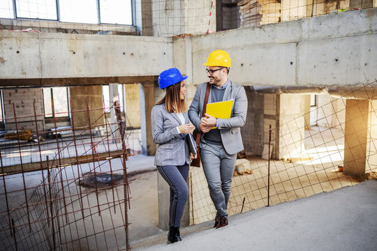 Two smiling positive handsome colleagues with helmets on heads looking at each other, talking and climbing the stairs in old building they want to rebuilt.
