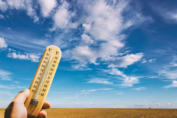 Thermometer in Hand with Clouds on Blue Sky and Field on Background