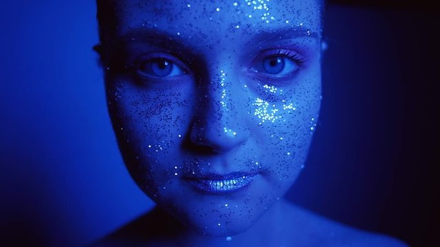 Neon light portrait. Skincare beauty. Woman with glitter face opening eyes slowly in blue glow.
