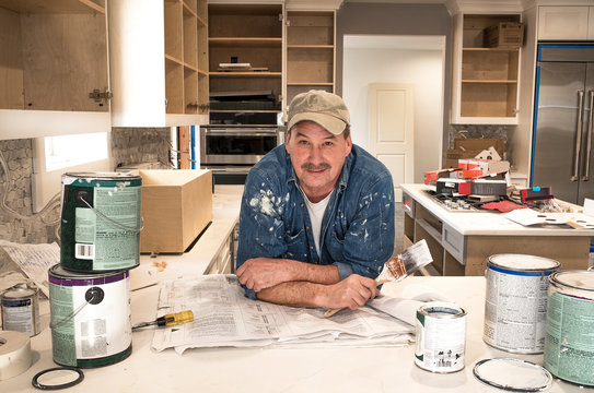 Male painter splattered with paint and holding a wet paint brush in messy home kitchen during residential home remodel with empty paint cans stacked around him, cabinet doors off and open