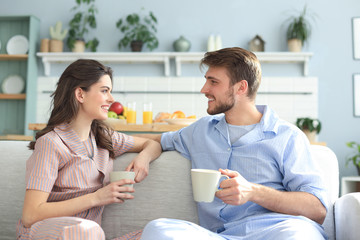 Beautiful young couple in pajamas is looking at each other and smiling on a sofa in the living room.