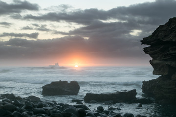 A boat in stormy seas at dawn with rocks in the foreground