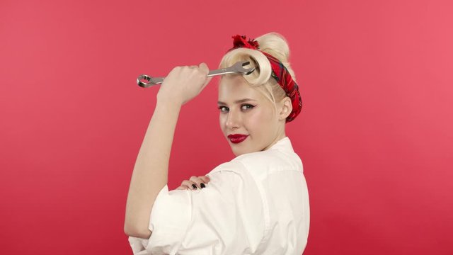 Pin up blonde woman holding wrench and touching biceps on pink background