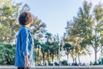 Afro brunette girl with blue denim shirt with her back to the camera looks to the other side of a park