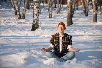 Ginger woman sitting in winter near tree. Young woman doing yoga lotus position in winter snow forest. Freedom and joyful concept.