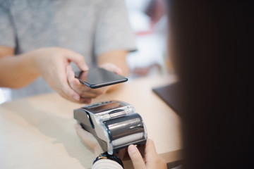 Hand of man customer using wireless or contactless payment of a smart phone. Young Asian cashier or seller are smiling to accept payment by nfc technology at retail shop. Contactless payment concept.