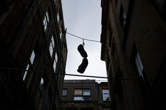 A silhouette image of shoes that are tied to each other by the shoe-strings, hangs from an electrical wire in an alley of the urban area of Glasgow, Scotland on a cloudy day. 
