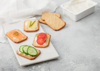 Various healthy crackers with salmon and cheese, tomato and cucumber on marble board on light kitchen table background with cream and kitchen towel