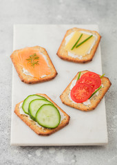 Various healthy crackers with salmon and cheese, tomato and cucumber on marble board on light kitchen table background.