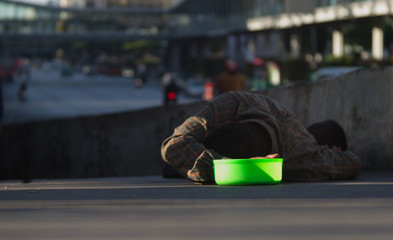 A beggar is laying down on a floor with his green plastic bowl