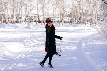 A young brunette in warm clothes with skates in her hands, against the backdrop of a city park full of snow. Winter vacation concept.