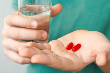 a young man holding two red capsules with medicine and a glass of water closeup; health care concept; additives, vitamins or painkiller
