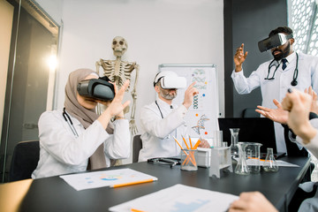 Group of multiethnic doctors scientists, African and Caucasian men, Muslim woman, wearing vr glasses working with 3d virtual content at modern workplace. Human skeleton on the background