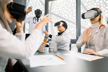 Team of multiethnical doctors, lab workers, scientists, Cacasian and Muslim women, African and Asian men, using virtual reality glasses for research and imagination sitting at the table in laboratory