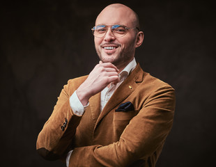 Fototapeta na wymiar Successfull and impressed attractive adult bald smiling businessman posing for camera in a dark studio wearing stylish mustard color velvet jacket, white shirt and glasses