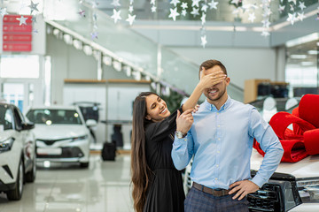 lovely young caucasian couple stand with car in dealership, new auto with red bow in the background. lady closed man's eyes goinh to make surprise
