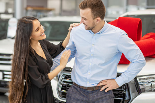 Beautiful Married Couple With New Automobile In Representative Dealership. Happy Caucasian Man And Woman After Purchase Of Car. New Car With Red Bow As A Gift In The Background