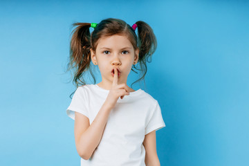 Fototapeta premium portrait of young caucasian serious kid girl in white t-shirt, stand with pigtails isolated over blue background, little child want people to be quiet. show tss gesture