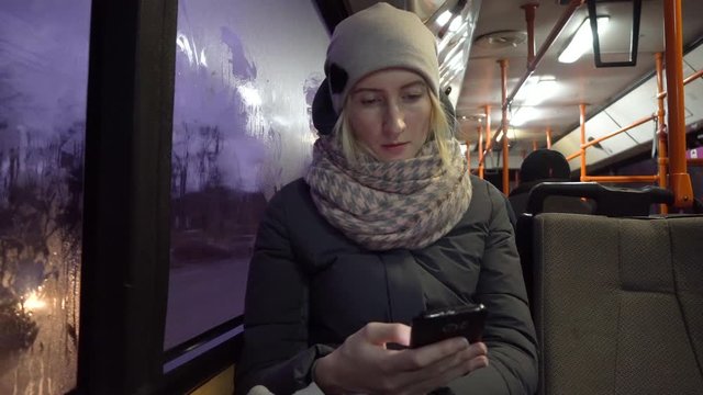 Attractive young woman using a smartphone while riding on public transport and looking out the window. Night time city of lights background.