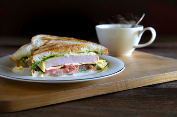Sandwich cheese and thick ham with veg. in white dish and black coffee all put on wooden chopping board