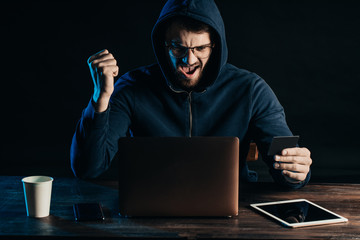 happy hacker male sit in hood of blue pullover, male sit cheerfully look at screen of laptop after...