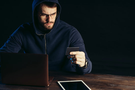 young caucasian man hacking bank card using computer system, male wearing pullover, sit in the hood at the table, seriously look at numbers of card and entering the code at laptop. cyberattack concept