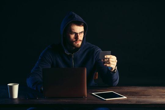 seriously looking man hacker engaged in illegal hacking, use laptop and tablet for hacking. male wearing pullover, sit in the hood isolated over black background. cyberattack, hacking, crime concept