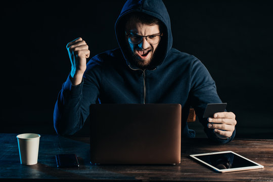 happy hacker male sit in hood of blue pullover, male sit cheerfully look at screen of laptop after successful hacking, hold bank card in hand, use laptop and tablet