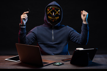 anonymous hacker in mask after successful getting access to bank card, raised hands up, hold bank...