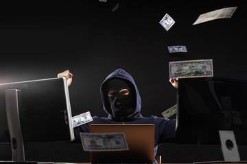 pleased young anonymous hacker man sit in mask in space full of money, dollar bills fly around the...