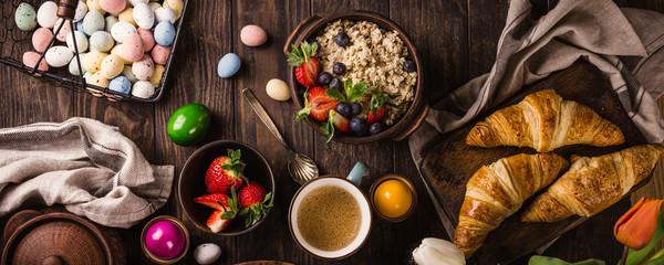 Rustic easter breakfast flat lay with eggs bagels, tulips, croissants, egg, oatmeal with berries, colored quail eggs and spring holidays decorations. Top view, banner.