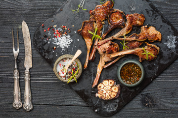 Grilling lamb chops with salt, pepper and dry herbs on dark stone plate on black old rustic wooden table, flat lay 