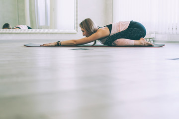 Young woman doing stretching exercise by mirror in bright yoga class room