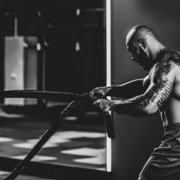 Side view of powerful athlete doing exercises with rope in modern gym, dramatic black and white image