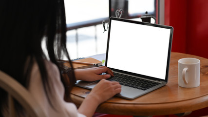 Woman working as website administrator typing on keyboard of white blank screen computer laptop while sitting at the modern wooden table with comfortable living room as background.