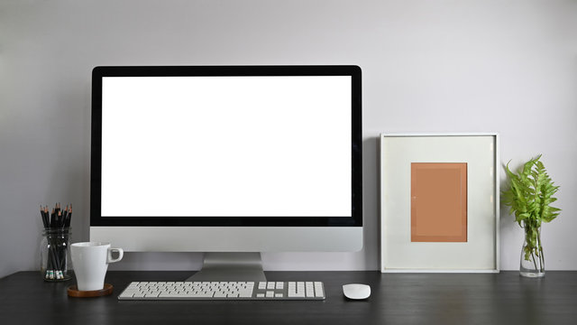 Personal workspace computer with blank screen, pencil holder, picture frame, potted plant, and coffee cup putting together on modern wooden table with white wall as background.