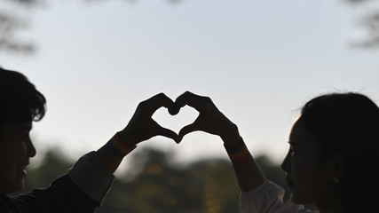 Silhouette image of young LGBT lesbian couple making a heart sign by hands while standing together at natural outdoor/parks with blurred forest and evening sky as background. - obrazy, fototapety, plakaty