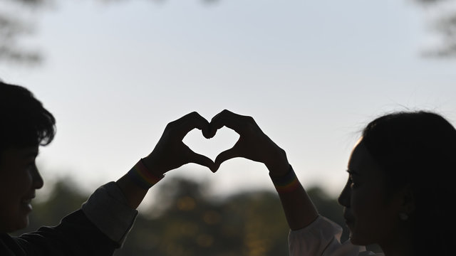 Silhouette image of young LGBT lesbian couple making a heart sign by hands while standing together at natural outdoor/parks with blurred forest and evening sky as background.