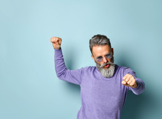 Gray-haired aged male in purple sweater, sunglasses. Dancing with clenched fists while posing on blue studio background. Close up