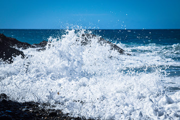 Large wave breaking on black rocks at Ajuy beach in Fuerteventura. Lots of foam and spray. Atlantic Ocean beyond with horizon and blue sky.