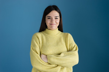 Portrait of caucasian girl young woman posing in front of blue background wall wearing yellow sweater crossed arms smiling black hair looking to the camera