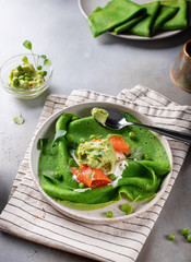 green spinach pancakes or crepes with salmon and mashed avocado on a ceramic plate. gray background, vertical image