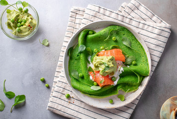 green spinach pancakes (crepes, blini) with salmon, mashed avocado, peas, arugula. gray background,...