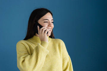 Portrait of caucasian girl young woman posing in front of blue background wall wearing yellow sweater using smart phone making a phone call talking smiling